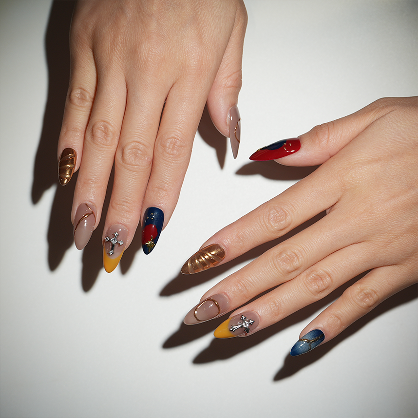 Hand showcasing long stiletto press-on nails in bold red, blue, and gold designs with cross charms and metallic accents for an elegant statement.