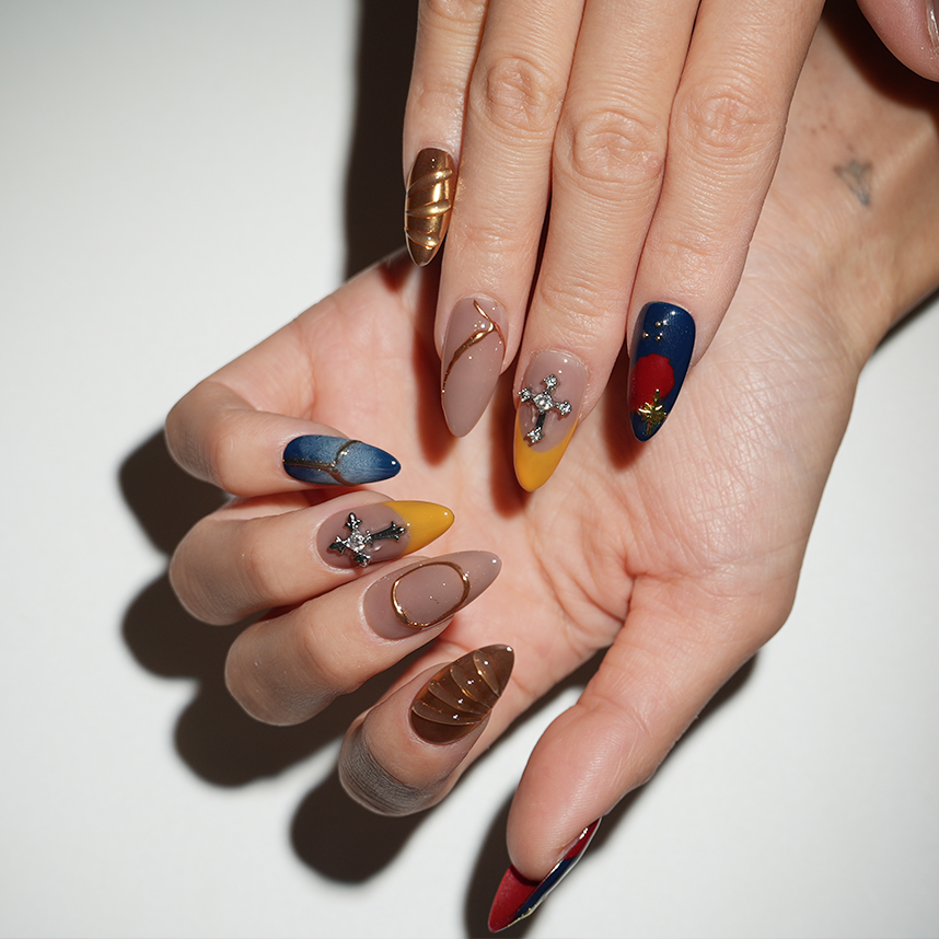 Close-up of hands wearing artistic press-on nails featuring red, blue, and gold tones with cross details and geometric metallic designs, perfect for a modern luxe look.